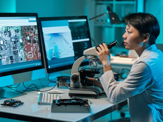 Young serious female scientist with microscope looking at computer screen while sitting by workplace
